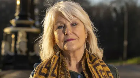 Liam McBurney/PA Wire Margaret McGuckin, a woman with shoulder-length blonde hair, pictured in the grounds of Stormont on a sunny day in summer 2023. She is leaning one elbow on a long stone wall. She is wearing a black leather jacket and a large brown and tan patterned scarf. 