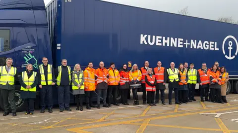 People in high visibility jackets stand in front of a blue lorry
