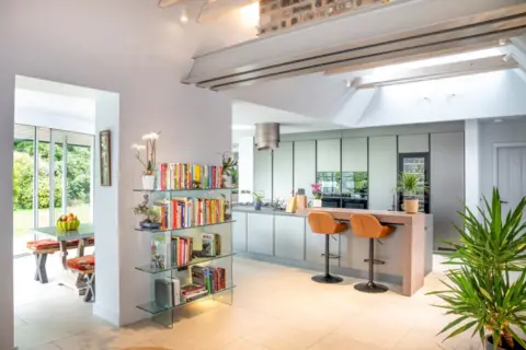 IWC Media Inside Art Deco Villa. The kitchen is modern with a long kitchen island, bar stools, house plants and a glass book case. The walls are white and the room is bright.