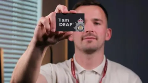 Matthew Wheeler is holding up a card that says "I am DEAF" on it and a logo of Cambridgeshire Police. He has short black hair and stubble and is wearing a white polo neck shirt, with a red lanyard around his neck.