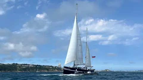 BBC A large sailing boat with white sails in the sea with the coast of Guernsey in the background, blue skies with grey and white clouds
