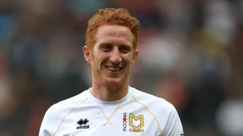 Dean Lewington is on a football pitch walking towards the camera. He has short ginger hair and a short bead. He is wearing a white football shirt with the MK Dons insignia on it.