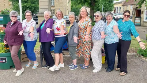 Donna Anderson A group of nine women, all in a row, standing in a market square, they all have their arms out. They are smiling. Some have their legs kicking out.