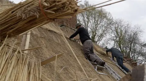 Jamie Niblock/BBC Two thatchers work on a roof. They rest on step ladders. Piles of straw can be seen near them. They are wearing long-sleeved tops, combat trousers and boots. There are trees in the background.