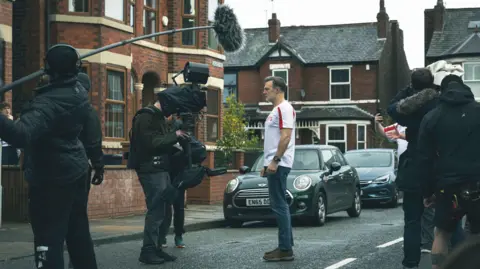 Channel 4 A wide shot of a film crew filming a scene with David Morrisey, who is wearing jeans and a football top and staring blankly into a camera