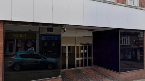 An empty shop. The windows are blacked out and there are two sets of doors in the middle, one of which is automatic. A lone CCTV camera points down towards the entrance.