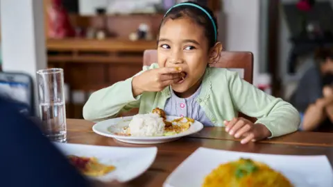 Getty Images Cute girl looking away while eating food at home. Female is having lunch at dining table. She is in casuals.