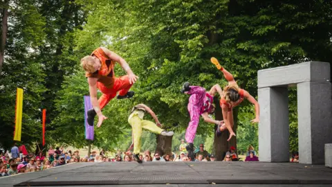 Becca Hicks/Greenbelt A small outdoor stage with four circus performers leaping and twisting in the air, wearing brightly coloured outfits. You can see the tops of the crowd watching. There are tall leafy trees behind.