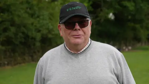 Shaun Whitmore/BBC A man wearing a black cap, sunglasses and a grey sweatshirt smiles at the camera. He is standing outside on a grassy area and there are large green trees behind him. 