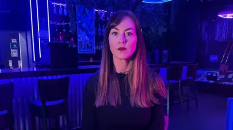 A woman with long brown hair stood in a nigtclub setting. The club is dark and has blue strobe lighting in the background.