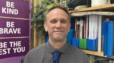 A man in a shirt and blue tie stood in front of bookshelves.