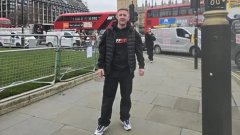 Gary Quinn Gary Quinn, a painter and decorator, stands on pavement in central London. with traders driving in vans behind him at a rally. Three red London buses are also behind him.