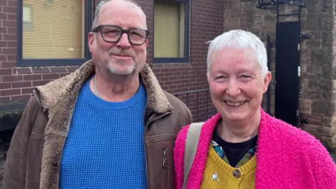 Christopher and Denise are both elderly. Denise has short grey hair and Christopher is bald and is wearing black glasses. They are both smiling at the camera. 