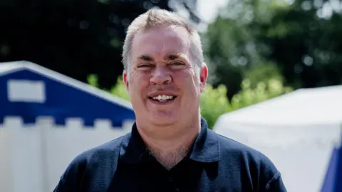 StreetGames Mark Lawrie, a man with grey hair and wearing a black polo shirt, smiling at the camera with trees behind him.