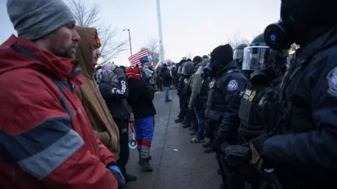 Manifestantes da Reuters ficam em frente à Alfândega e Proteção de Fronteiras dos EUA (CBP) e outras autoridades policiais perto do Edifício Federal Bishop Henry Whipple. Um homem está vestindo uma jaqueta vermelha, o outro está vestindo uma jaqueta marrom e um está usando capacetes pretos e pretos contra os oficiais de linha.
