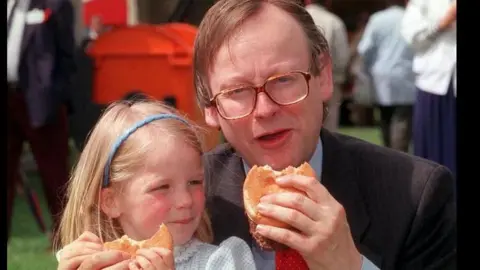 PA Media John Gummer wearing glasses, a dark suit jacket, a light-coloured shirt and a red tie is sitting outdoors holding a partially eaten burger. Beside him, a child wearing a light-coloured top and a blue headband is also holding a partially eaten burger. In the background are several out-of-focus people, a large orange bin and market or event-style tents. The photograph appears to have been taken at an outdoor public event during daylight.
