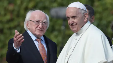 Danny Lawson/Pa Wire Michael D Higgins had white hair that is bald at the top. It wears a dark suit, a light blue shirt, an orange tie and glasses. He is holding a hand towards the camera. Beside Pope Francis, who is smiling at something out of the camera. Wear a white dress and a piece of head.
