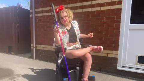 A woman sitting in a wheelchair holding a mobility stick in her right hand and a pink party blower in her mouth.