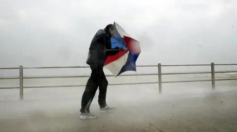 A man walking along a promenade, struggling against the wind. He is holding an umbrella in front of his face, using it as a shield against the wind. The red, white and blue umbrella is only half open. There is mist from the spray from the sea and rain around the man. He is wearing a black coat and black and grey tracksuit bottoms and trainers. 