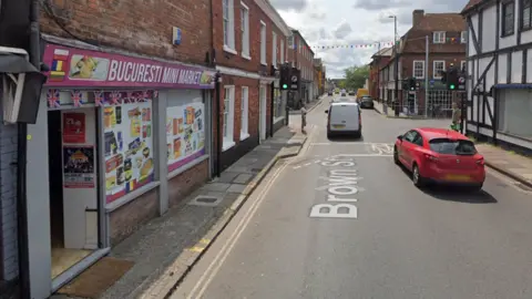Google Google maps image of a street in Salisbury. Bucuresti Mini Market is on the left - it has union jack bunting and a pink storefront. Pictures of various food and drink products can be seen on the windows. Traffic lights and other buildings can be seen along the street. 