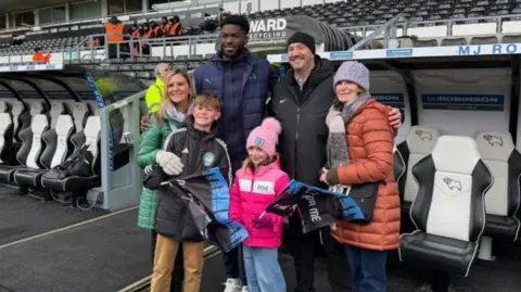 BBC Derby County player Patrick Agyemang flanked by two women, a man and two children. They are wearing thick coats and hats. They are stood in front of the dugouts pitch side.