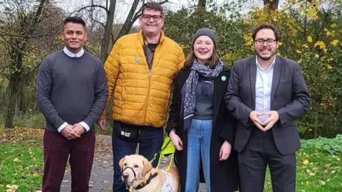 (Left to right) Khaled Musharraf, Alistair Chisholm, Sarah Peters, and Nick Hartley are standing in a leafy park and all smiling at the camera.