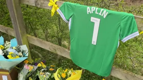 A green shirt with Amelia's name and the number 1 hangs from a fence whilst flowers lie in front of it.