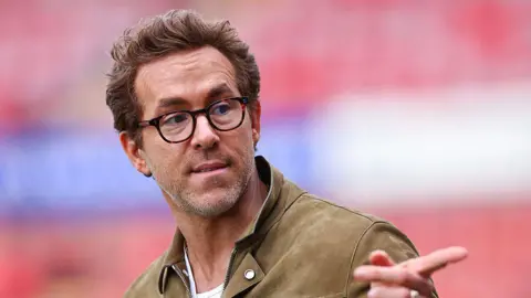 Ryan Reynolds, wearing glasses and a brown jacket, pointing a finger