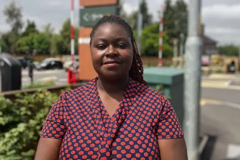 Owen Sennitt/BBC Makala Zulu, wearing a red and blue patterned top, looks towards the camera in front of the entrance to a McDonald's restaurant in Sprowston, Norwich