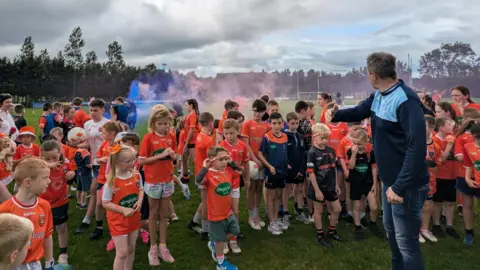 BBC Man with his back to camera waving to a crowd of children in orange jerseys to direct them. They are standing on a GAA pitch and there is flare smoke in the background 