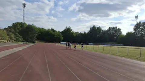 Tom Grantham A group of runners on an athletics track sprint on the inside lanes next to a grass field