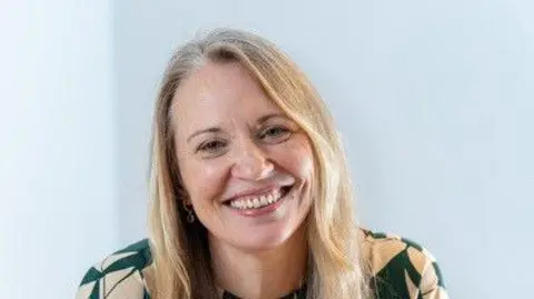 Leona Deakin A blonde woman smiles at the camera. She is wearing a green and white top and is sitting in front of a blue background.
