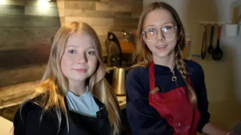 Two young people, Molly and Sinead, are pictured in a modern kitchen setting, standing side by side near a counter top. Both are wearing aprons. Molly on the left has long blonde hair, a light blue T-shirt and a black apron. She is smiling. Sinead has long brown hair, one length in a plait, and is wearing glasses and a red apron. Behind them, there are stainless steel appliances and utensils hanging in a contemporary-looking cooking psace.

