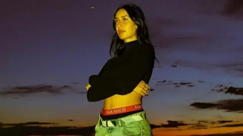 Julia Wolf Julia Wolf is pictured against a dramatic sunset. She is wearing a black crop top, with her arms folded across her chest as she stares into the distance.