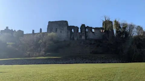 A general view of Cockermouth Castle from the footpath along the River Derwent. The large structure has missing windows and appears to be like a ruin on the right hand-side, while it looks more like a complete structure on the left. The castle overlooks the river and is in a slightly raised position from the riverbank.