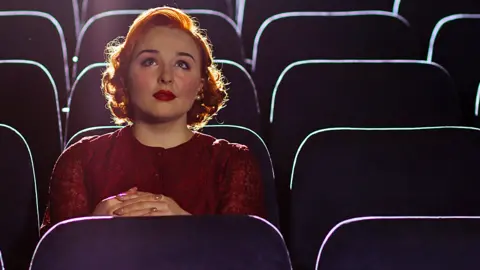 Woman sits alone among rows of empty chairs in auditorium