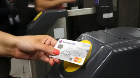 A Barclaycard credit card being placed on a contactless card reader at a train station ticket barrier.