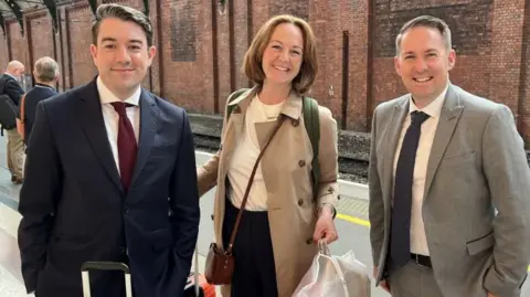 BBC The three new MPs stand on a platform at a train station smiling at the camera