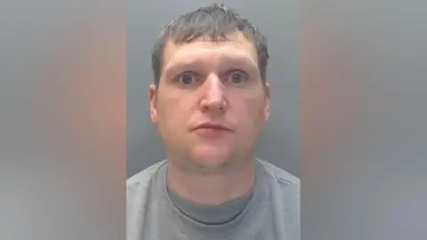 Durham Police Mugshot of Stockton. He is wearing a grey t-shirt and jumper and has short brown hair.