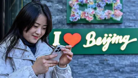 Getty Image A Chinese Woman checks her phone, reads with a sign in the background "Eye Heart Beijing"