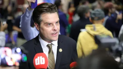 Getty Images Matt Gaetz, with his short brown hair combed back, wearing a suit with a striped red tie and golden lapel pin, takes questions from reporters around him at the ABC News presidential debate in September