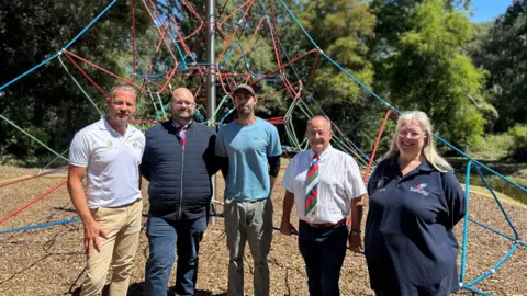 Some of the members of wooden spoon stood in a line in front of the spiders web climbing frame 