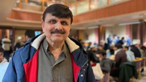 Sarfaraz smiles at the camera while people eat their food behind him. He is wearing a pale grey shirt with a blue coat on. He has short black hair and a moustache.