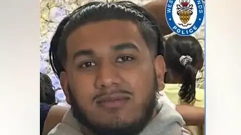 West Midlands Police A man with a short black beard and think moustache smiling and looking into the camera. There is a lilac flower wall behind him and his is wearing a thin, black headband to hold his hair out of his face. 