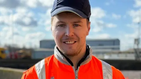 A man wearing an orange reflective jacket and a cap stands outside. Water and blue skies with clouds can be seen behind him.
