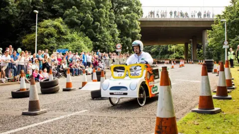 A person in a handmade white kart, wearing a white helmet and suit. The vehicle is on a track lined with orange cones and tires, with a large crowd of spectators and a bridge in the background.