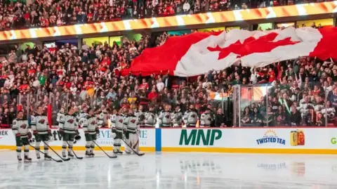 The Canadian flag is held up over spectators as the national anthem is sung prior to the start of a game between the Minnesota Wild and the Ottawa Senators at the Canadian Tire Centre in Ottowa. Ice hockey players are lined up on the rink holding their sticks.