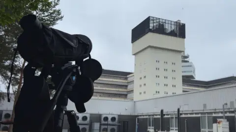 Stuart Howells/BBC A monocular on a tripod trained towards and industrial building with a large white tower