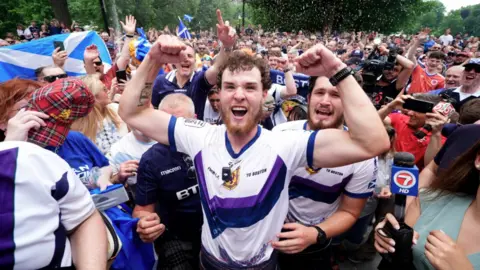 PA Media Crowd of sports fans in a park waving Scottish flags, with a central person in a white and purple jersey raising both fists while surrounded by reporters and onlookers.