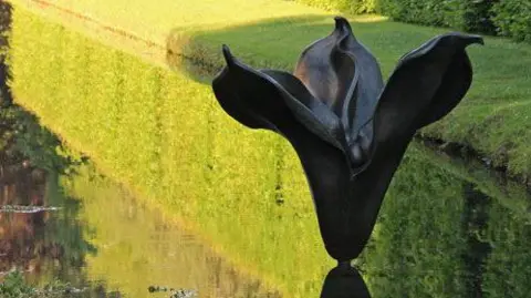 A brown, bronze sculpture called La Promesse, sitting in water, surrounded by grass. The artwork is based on the seed pod of the Iris Foetidissima plant, and is dark colour. A reflect of the work is on the water. 